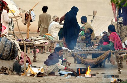 A nomadic woman cooks over an open flame as families rest on cots in a makeshift shelter, demonstrating the resilience of displaced people in difficult circumstances